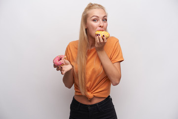 Excited young pretty long haired blonde woman looking poisitively at camera with raised eyebrows and tasting donats in her hands, wearing casual clothes over white background