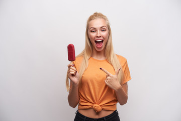 Overjoyed lovely young blonde woman with casual hairstyle pointing with forefinger on berry ice-cream in her hand, looking joyfully to camera with wide mouth opened, isolated over white background