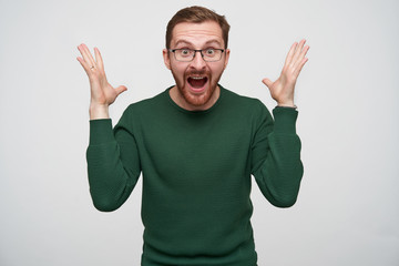 Overjoyed young pretty brunette bearded male in glasses raising happily hands and looking at camera with wide eyes and mouth opened, standing against white background