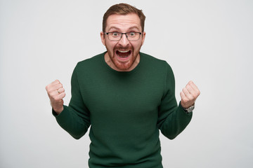 Fototapeta premium Emotional young handsome bearded male with brown short hair raising fists and screaming loud with wide eyes and mouth opened, isolated over white background