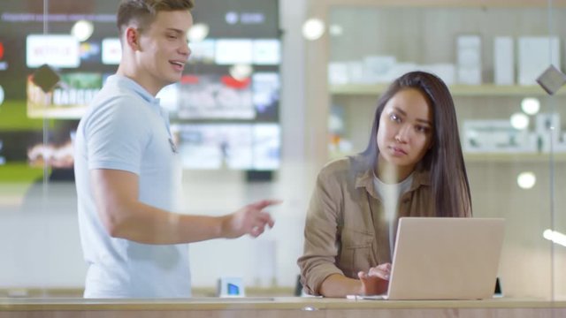 Young Asian woman testing laptop computer at gadget store, friendly male shop assistant approaching and talking to herYoung Asian woman testing laptop computer at gadget store, friendly male shop assi