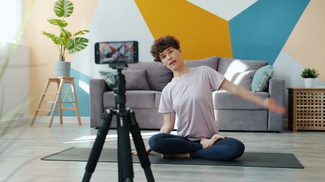 Smiling yoga instructor is recording tutorial at home vlogging using smartphone camera sitting on mat stretching body bending sidewards. Vlog and gadgets concept.