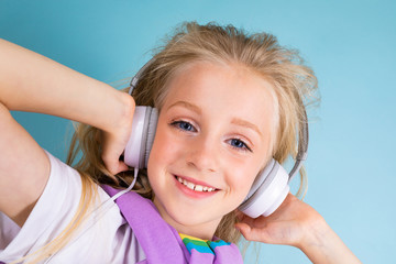 portrait of a beautiful blond charming young cute child in a casual look with white headphones listening to the music and on a light blue background
