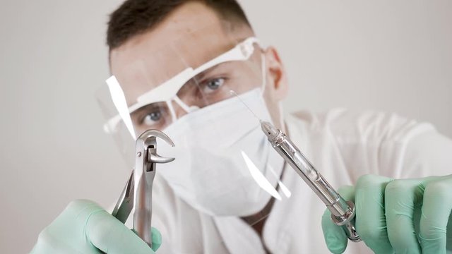 A caucasian male dentist in a mask and glasses releases air bubbles from a syringe with a preparation and holds dental forceps in his hand to remove teeth. Concept of dental care and dental treatment.