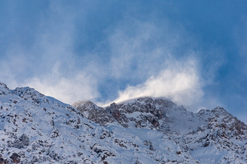 Snow dust in the sun in the air on the top of a mountain against a blue sky. Strong wind on top of the mountain. The wind tears the snow cover from the top of mount. Mountain peak in the cloud.