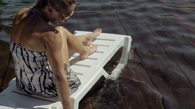 Woman lie on a sunbed in sunglasses and a boho silk shawl. Girl rest on a flood wood underwater pier. The pavement is covered with water in the lake. In the background are mountain and a forest.