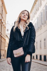 street style portrait of european attractive blonde woman wearing leather trousers and jacket. crossing the street and smiling at camera. fashion outfit details perfect for autumn fall winter