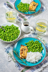 Baked red fish in a sauce with a sprig of rosemary and a slice of lemon on a bright plate with green peas and a side dish of rice, a glass of water with lemon on a light gray background