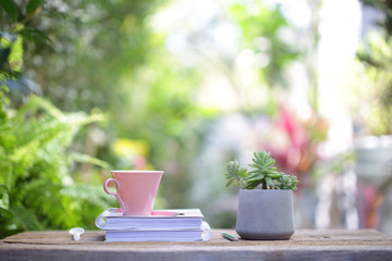 pink coffee cup with small Succulent plant in old grey concrete pot with white thick book on wooden table at outdoor with nature bokeh background