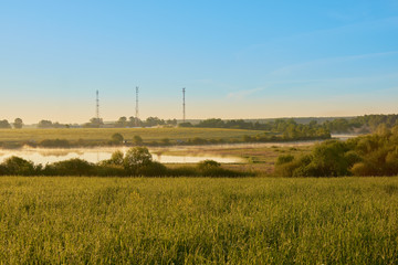 dawn with a view of the field and the river with three cell towers on the shore with clouds in the sky.