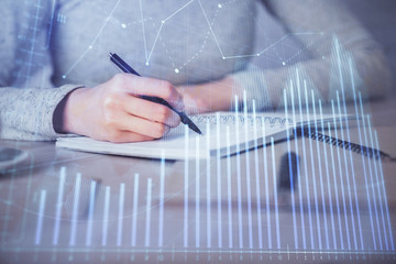 Hands writing information about stock market in notepad. Forex chart holograms in front. Concept of...