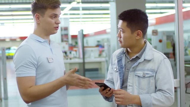Medium shot of young Asian man trying to choose smartphone at gadget store, shop assistant helping him