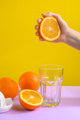 Juicy fresh orange in female hands hovers over a glass. Against a bright background, purple-yellow. Hard creative light, vertical photo.