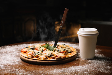 Hot large pizza and a glass of coffee tea pizza composition with melting cheese chicken tomatoes mushrooms steam smoke on a black background