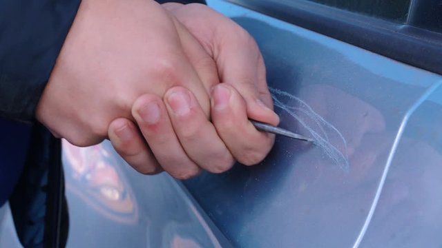 Bad guy scratching the car door with a screwdriver in the parking lot on the street. Damage of property from revenge for treason or betrayal, or threat. Auto insurance fraud or vandalism.