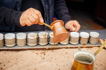 Closeup man brews turkish coffee in the sand. Pours coffee into traditional coffee cups.