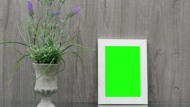 white photo frame with green chroma-key picture stands near purple flowers of grey pot plant on wooden background zoom in