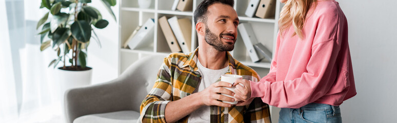 panoramic shot of woman giving cup of tea to bearded man
