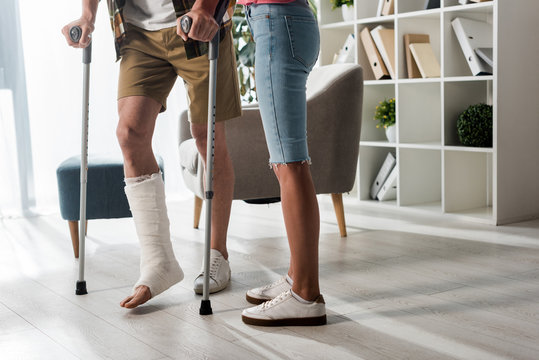 cropped view of woman standing near man holding crutches at home