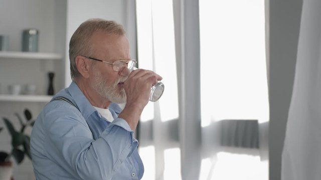 sick mature man with a gray beard retirement takes a pill with water for health and feels better at home near window