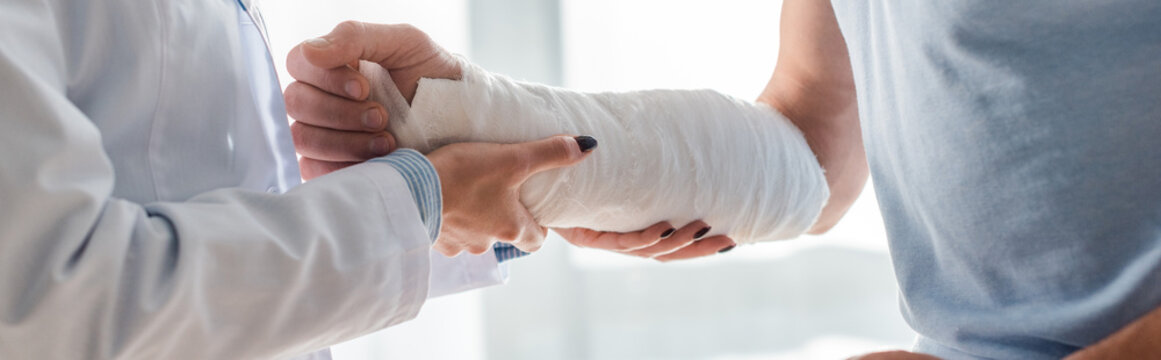 panoramic shot of orthopedist touching injured hand of man