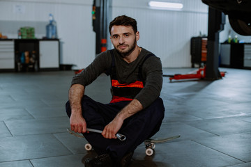 Fototapeta na wymiar A young mechanic is sitting on a skateboard in a car service after finishing work smiling