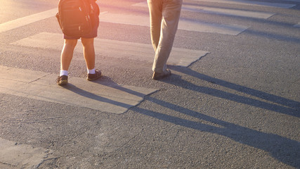 Low section and rear view of Asian elementary student boy in uniform with his parent are walking across the zebra crossway with flare light and shadow on asphalt road surface in evening time