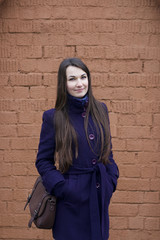 Girl in a purple coat posing on a background of a brown brick wall. Holds hands in pockets.