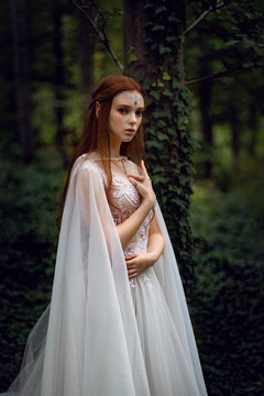 Attractive ginger elf woman in white dress and cape posing in green garden 