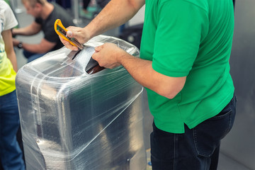Worker wrapping suitcase with transparent protection film atairport deparure terminal before...