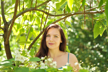 Beautiful brunette woman with long hair and blue eyes walking in the blooming park at spring in sunny day with bright sunshines and green trees