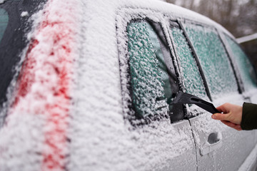 Cleaning the side car windows of snow with ice scraper before the trip. Man removes ice from car windows. Male hand cleans car with special tool at snowy frosty winter day.