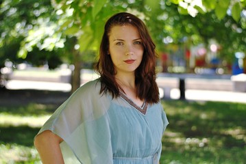 Beautiful brunette woman with long hair and blue eyes walking in the blooming park at spring in sunny day with bright sunshines and green trees