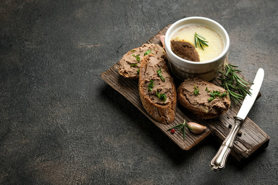 Fresh homemade chicken liver pate in ceramic bowl or ramekin and baguette slices with herbs and garlic on a dark background.