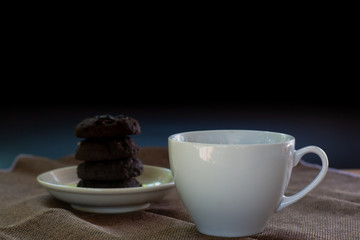 White ceramic cup of coffee serve with blurry dark chocolate cookies on black background.