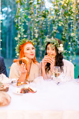 Two pretty hungry young bridesmaids in white dresses eating big burgers and smiling. Rustic wedding reception outdoors