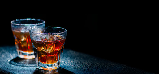 Two glass of cold alcohol cocktail drink on black background with copyspace. Whiskey with ice and cola on bar table.