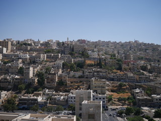 Cityscape of Amman, capitol of Jordan, grey panorama of a modern Arabic city with improvised houses on a hill between few green trees and a Roman theatre under the blue sky	