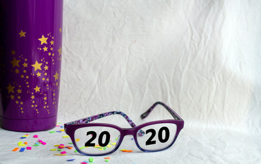 Obraz na płótnie Canvas Announcing 2020 in both vision and a new year with a party concept including a purple cup, stars, confetti, copy space and bokeh effect.