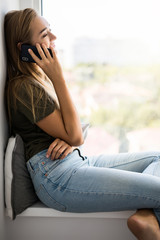 Obraz premium Beautiful young woman talking by mobile phone near window at home