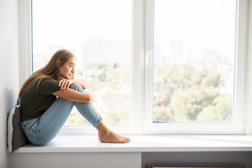 Sad depressed young woman having social problems sitting on windowsill - obrazy, fototapety, plakaty