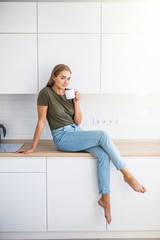 Young pretty woman holding cup with coffee or tea while sitting on counter in the kitchen