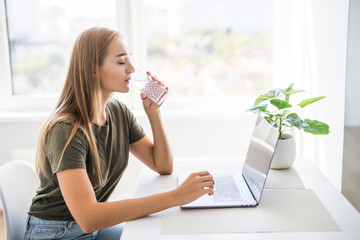 Woman drinking from water glass while typing at her laptop. Thirsty woman staying hydrated while working from home