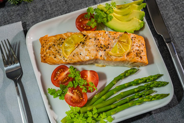 Baked salmon with lemon on a white plate with asparagus, avocado, tomatoes