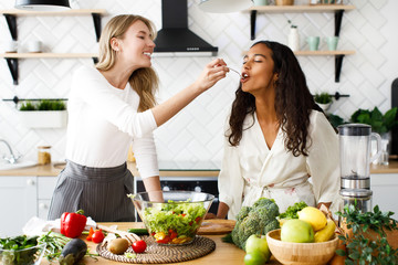 Attractive caucasian girl is feeding beautiful girl with fresh salad on  modern designed kitchen
