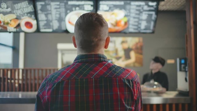 Back Shot Male Person Near Cash Desk In Fastfood Cafe. Man In Casual Wear Checkered Shirt Inside Food Restaurant Looking To Menu Board Choosing Dish To Eat