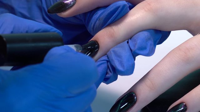 Closeup view of beautiful manicured nails of woman and professional manicurist applying transparent finish top polish laer of gel polish. Black design with shiny sparkling cat eye effect.