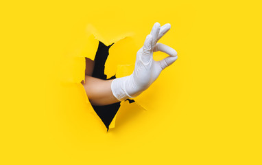 Doctor's hand in a white medical glove shows OK gesture. Yellow paper background, torn hole. Positive treatment concept. Copy space.