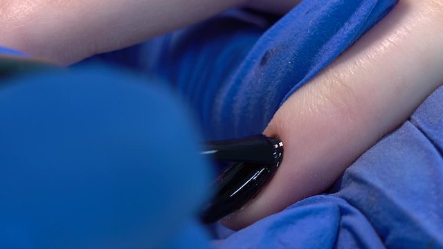 Closeup view of beautiful manicured nails of woman and professional manicurist applying transparent finish top polish laer of gel polish. Black design with shiny sparkling cat eye effect.