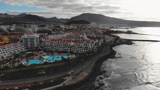Aerial view of Las Americas city, Tenerife, Canary islands. Cinematic 4K footage with black volcanic rocky coast, Atlantic ocean, waves, city modern villas, quay with people, mountains. Early morning